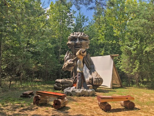 A large wooden sculpture of a lion-like creature sits near a campfire, surrounded by trees and a tent.