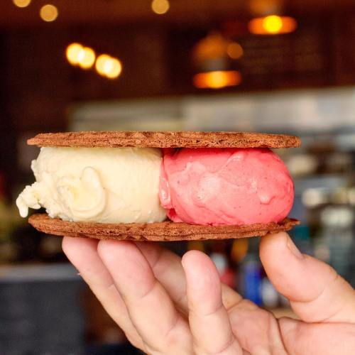 A hand holding a chocolate cookie sandwich with vanilla and strawberry ice cream inside.