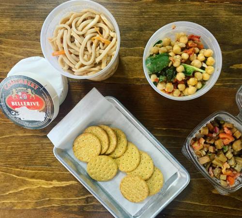 A wooden table with various food containers: noodles, chickpea salad, crackers, and a side dish.