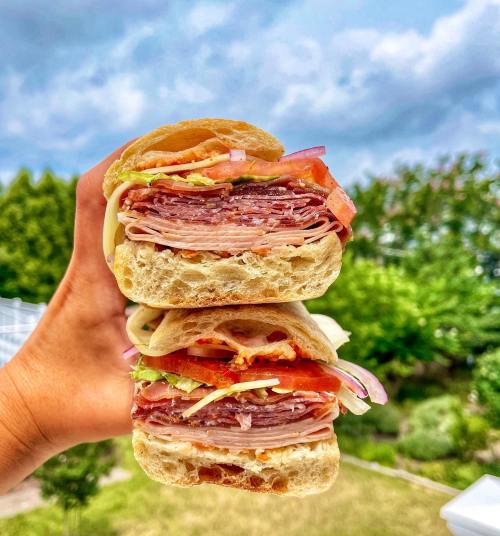 A hand holding a stacked sandwich filled with layers of meats, lettuce, tomatoes, and onions against a cloudy sky.