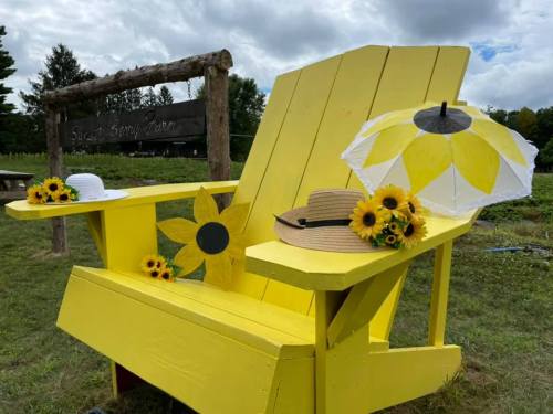 A bright yellow Adirondack chair adorned with sunflowers, a sun hat, and a sun umbrella in a field setting.