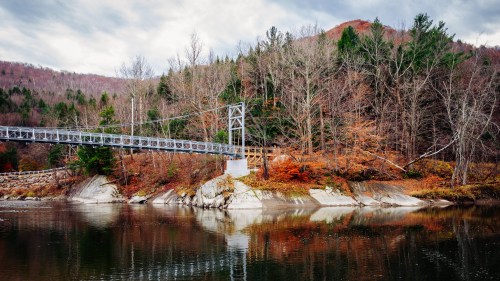 A suspension bridge spans a calm river, surrounded by autumn foliage and mountains under a cloudy sky.