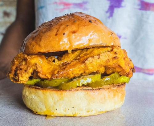 A close-up of a fried chicken sandwich with pickles on a soft bun, drizzled with sauce.