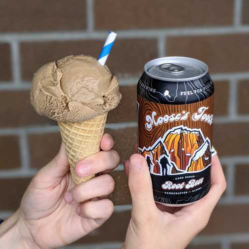 A hand holds a cone of chocolate ice cream and a can of Moose's Tooth Root Beer against a brick background.