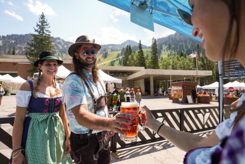 A man in traditional attire hands a large beer to a woman at an outdoor festival, with mountains in the background.