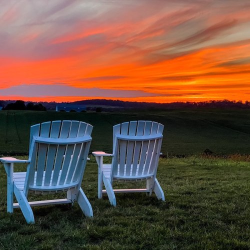 Two white adirondack chairs facing a vibrant sunset over rolling hills, with a colorful sky in shades of orange and purple.