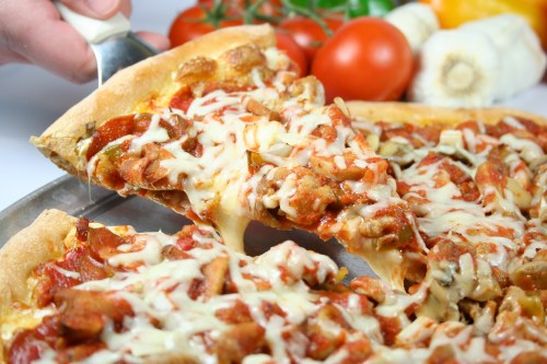 A hand lifts a slice of cheesy pizza topped with meat and vegetables, surrounded by fresh tomatoes and garlic.