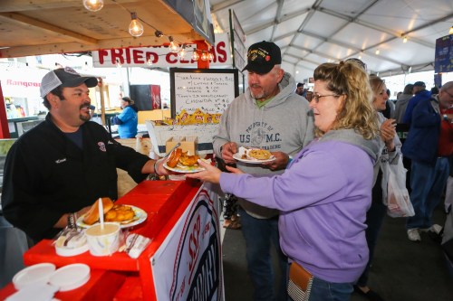 A vendor serves fried food to two customers at a bustling food festival under a tent.
