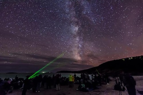 A crowd on a beach gazes at the Milky Way, with a green laser pointing towards the stars.