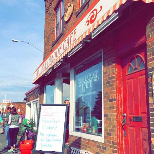 A cozy café with a red door and striped awning, featuring a sidewalk menu and people enjoying the outdoor space.