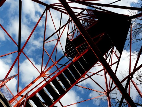 View looking up at a red metal tower structure with a staircase against a blue sky and scattered clouds.