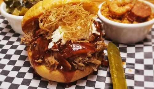 A pulled pork sandwich topped with crispy onions, served with sides of shrimp jambalaya and pickles on a checkered plate.