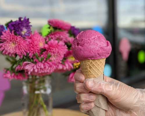 A hand in a glove holds a cone of vibrant purple ice cream, with a bouquet of pink flowers in the background.