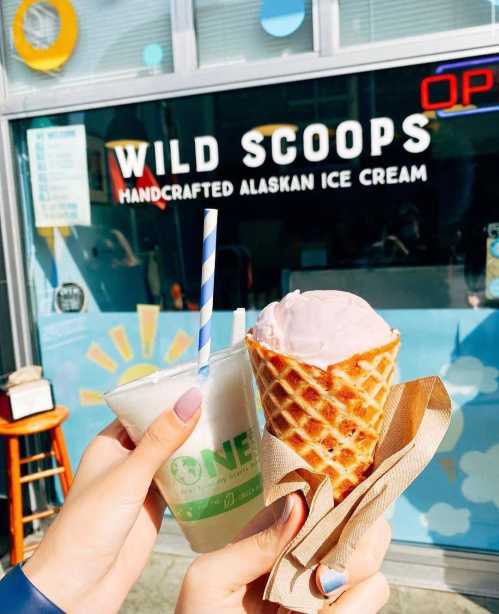 Two hands hold ice cream treats in front of a shop window that reads "Wild Scoops - Handcrafted Alaskan Ice Cream."