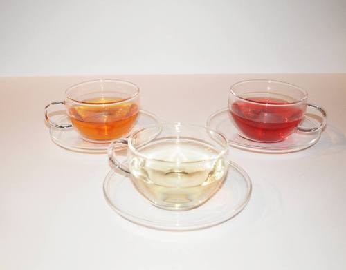 Three glass cups of tea: one amber, one clear, and one red, each on a matching saucer against a white background.