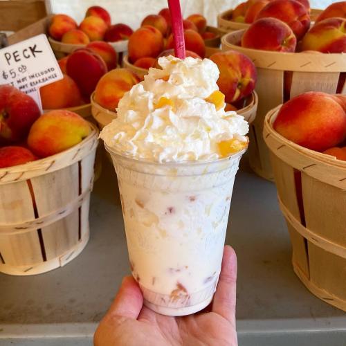 A hand holds a cup of creamy dessert topped with whipped cream, with baskets of fresh peaches in the background.