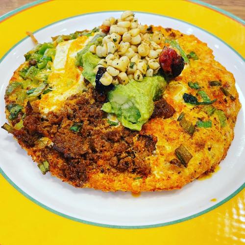A colorful plate featuring a round omelet topped with guacamole, corn, and herbs on a yellow rimmed plate.