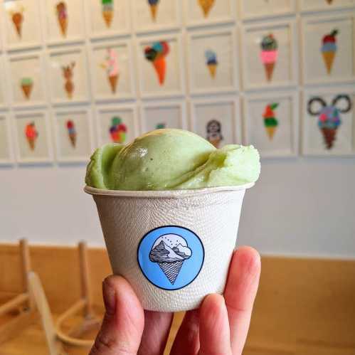 A hand holding a cup of green ice cream, with a colorful ice cream cone wall in the background.