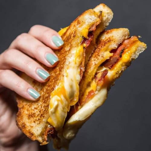 A hand holding a gooey grilled cheese sandwich with layers of melted cheese, bacon, and tomato, on toasted bread.