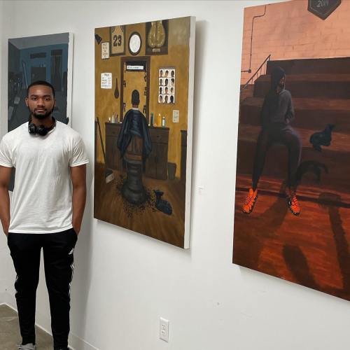 A man stands beside two paintings in an art gallery, one featuring a barber's chair and the other depicting a figure in a hoodie.