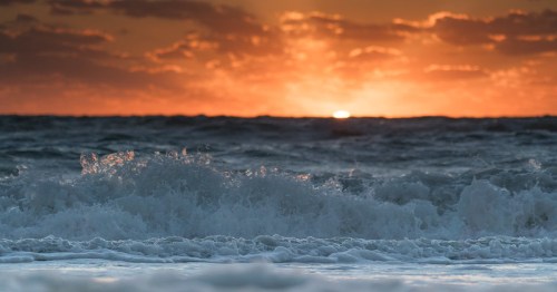 Waves crashing on a beach at sunset, with vibrant orange and yellow hues in the sky.