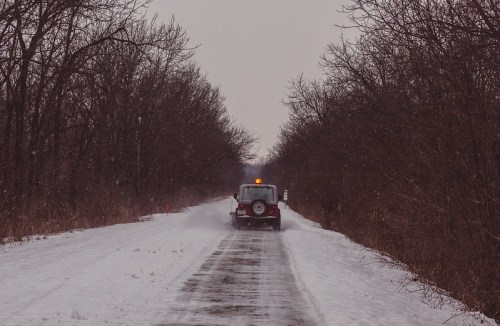 A snow-covered path with a vehicle driving through light snowfall, surrounded by bare trees.