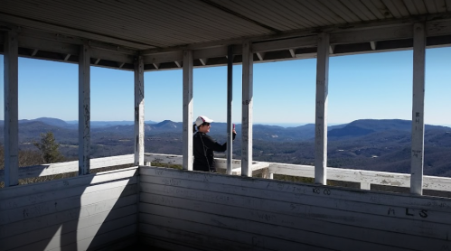 A person stands inside a lookout tower, gazing at a panoramic view of mountains and blue sky.