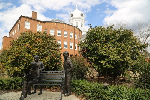 A bronze statue of children on a bench, surrounded by greenery, with a historic building and cloudy sky in the background.