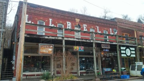 Historic brick building with a faded sign, featuring storefronts and a wooden balcony in a quaint town setting.