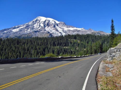 A scenic view of a winding road with a snow-capped mountain and dense forest under a clear blue sky.