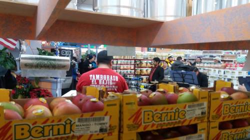 A grocery store scene with colorful fruit displays and shoppers in the background, including a musician performing.