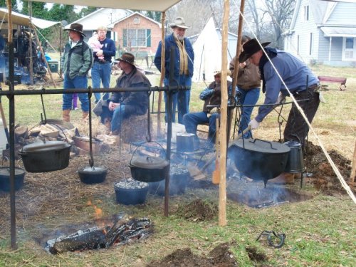 A group of people in cowboy attire gather around a campfire, with pots cooking over an open flame under a tent.