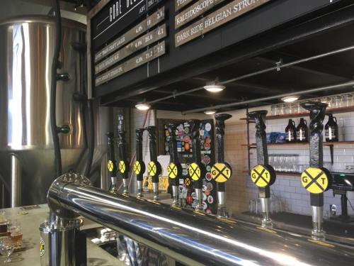A row of beer taps with yellow handles in a brewery, featuring a large brewing tank and a menu board in the background.