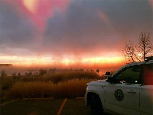 A white vehicle parked near a misty lake at sunrise, with colorful clouds and silhouettes of trees in the background.