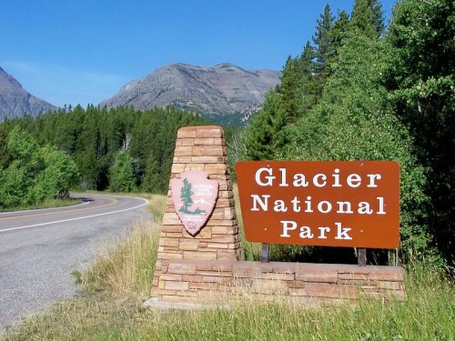 Sign for Glacier National Park beside a winding road, surrounded by trees and mountains in the background.