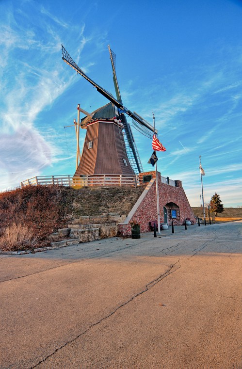 A historic windmill stands against a blue sky, surrounded by flags and a paved pathway.