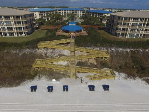 Aerial view of a beach with wooden stairs leading to the shore, surrounded by beach chairs and resort buildings.