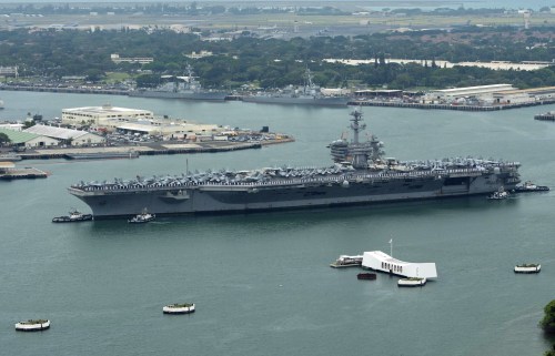 Aerial view of a large aircraft carrier docked in a harbor, surrounded by smaller boats and buildings in the background.