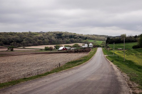 A rural road leads through fields to a farm, with rolling hills and overcast skies in the background.