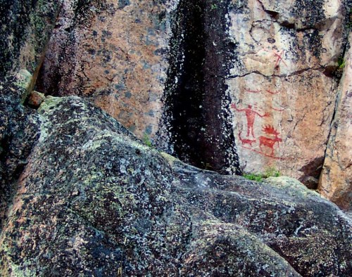 Ancient rock art depicting a figure and animal on a textured stone surface, surrounded by moss and lichen.