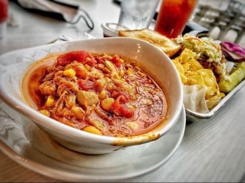 A bowl of hearty chili with tomatoes and corn, served alongside a plate of sandwiches and pickles.