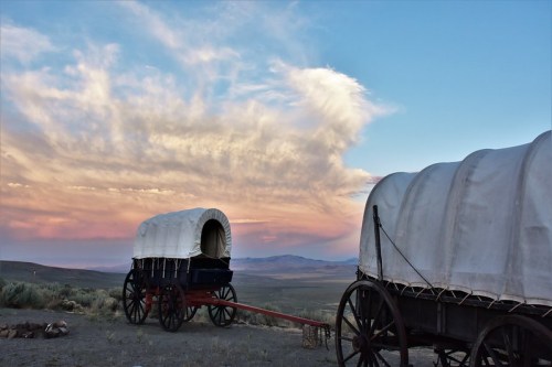 Two covered wagons sit on a dusty landscape under a colorful sunset sky with clouds.