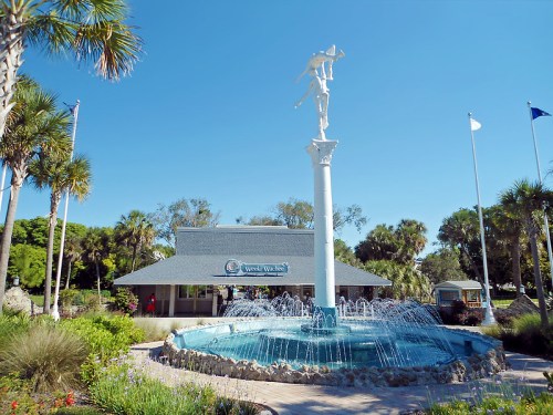 A fountain with a statue in front of a building surrounded by palm trees and greenery under a clear blue sky.