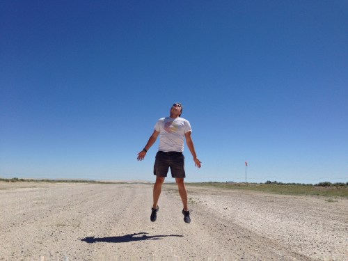 A person jumps joyfully on a dirt road under a clear blue sky, casting a shadow on the ground.