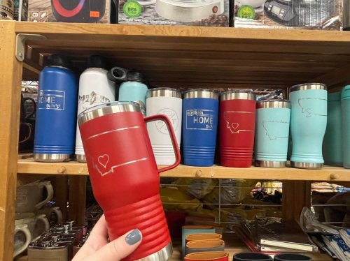 A hand holds a red travel mug among various colorful mugs on a wooden shelf.