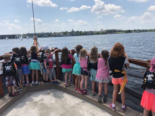 A group of children in pirate costumes looking out over a body of water from a boat on a sunny day.