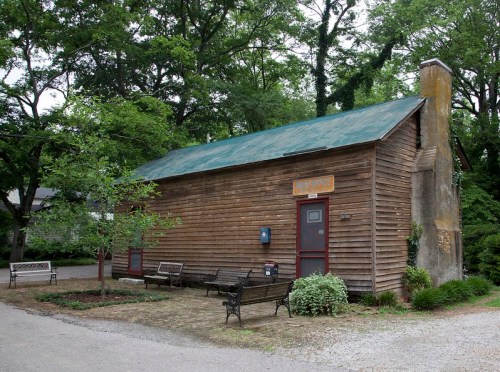 A small, rustic wooden building with a green roof, surrounded by trees and benches in a park-like setting.
