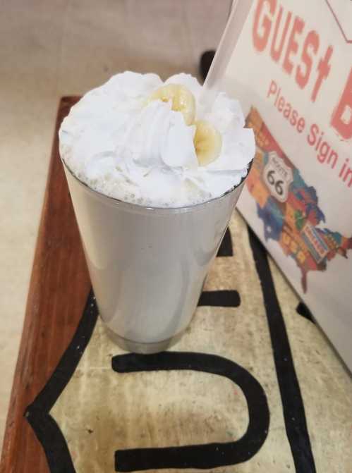 A tall glass filled with a creamy beverage topped with whipped cream and banana slices, placed on a wooden surface.