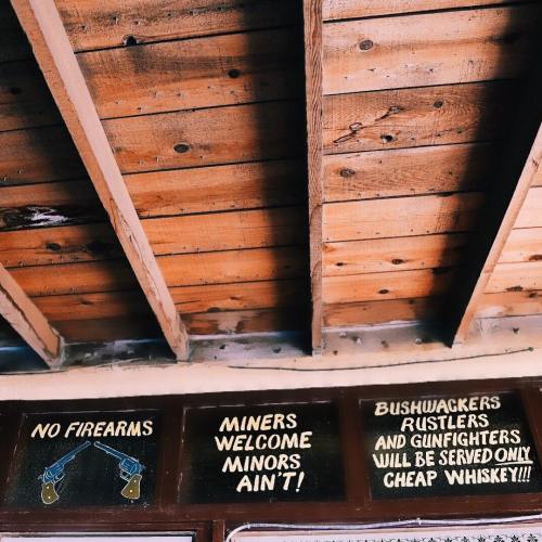 Sign on a wooden wall reads: "No firearms. Miners welcome, minors ain't! Bushwackers, rustlers, and gunfighters: cheap whiskey!"