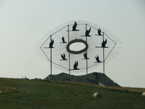 A large metal sculpture featuring birds in flight, set against a hazy sky, with a circular frame at its center.